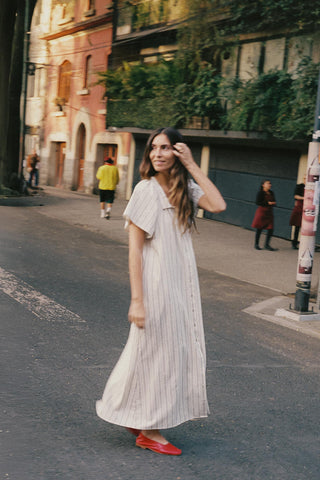 Woman in a light dress standing on a street with buildings and greenery in the background