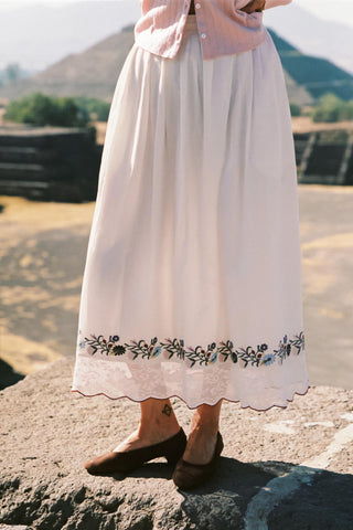 Person wearing a long white skirt with floral embroidery standing on a rock with a scenic background.