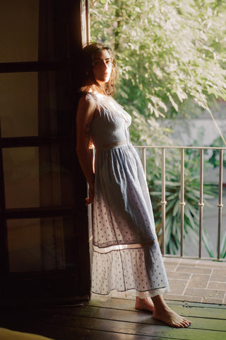 Woman in a light blue dress standing by a window with greenery outside