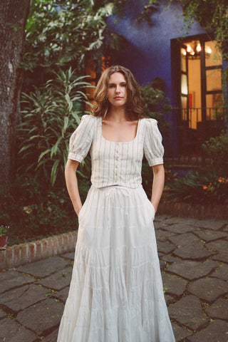 Woman in a striped top over a white skirt standing outdoors with greenery and a building in the background