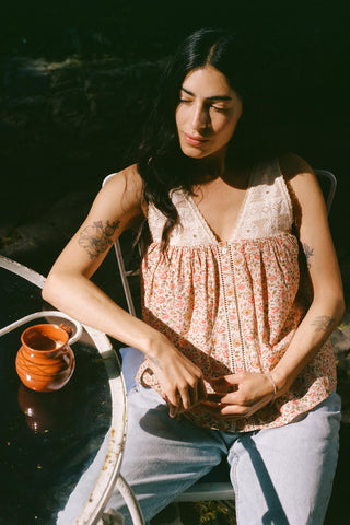 Woman sitting outdoors with a cup, wearing a patterned top and light pants.