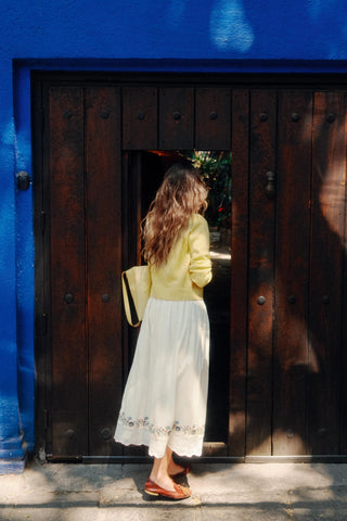 Woman in a yellow top and white skirt standing in front of a dark wooden door with a blue wall.