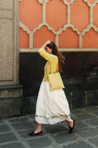 Woman in yellow sweater and white skirt walking past a decorative wall.