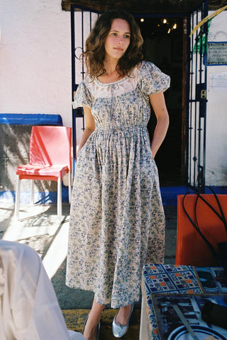 Woman in a floral dress standing in an outdoor setting with colorful chairs and a table.