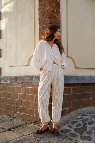 Woman in a white blouse and striped pants standing against a brick wall.