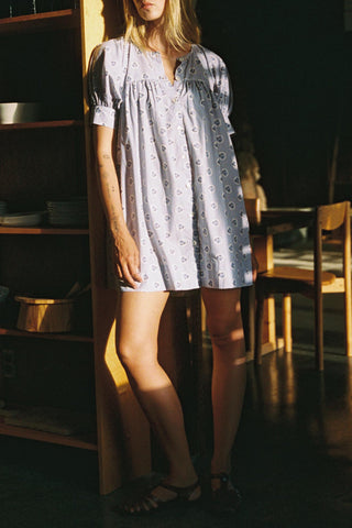 Woman wearing a light purple dress with white patterns in a dimly lit room.