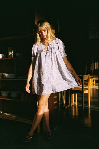 Woman in a light dress standing in a dimly lit room with shelves and chairs.