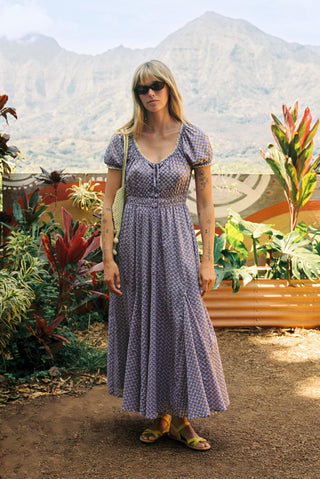 Woman in a long purple dress standing outdoors with mountains and plants in the background
