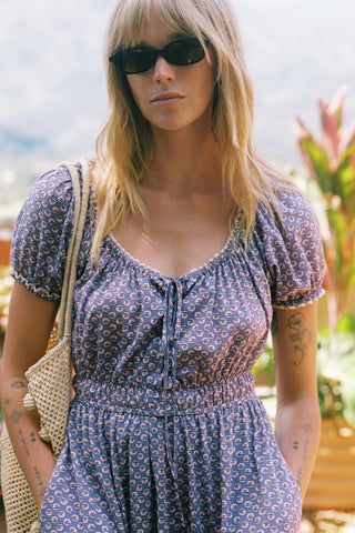 Woman wearing a purple floral dress with sunglasses and a beige bag outdoors.