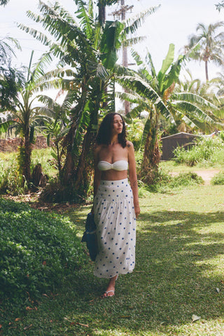 Woman in a white polka dot dress standing in a lush green garden with palm trees.