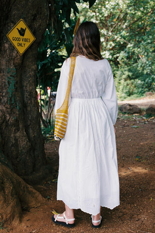 Woman in a white dress walking away from the camera in a forested area with a 'Good Vibes Only' sign on a tree.