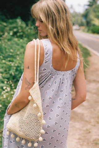 Woman in a light dress holding a beige woven bag with pom-poms outdoors.