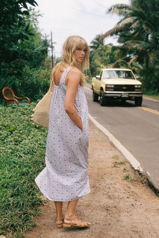 Woman in a white patterned dress standing on the side of a road with a truck in the background.