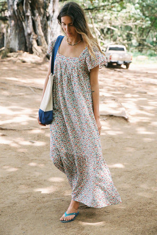 Woman in a floral dress walking on a dirt path with trees and a car in the background