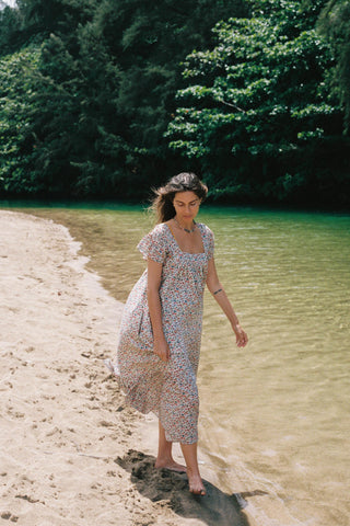 Woman in a floral dress standing on a sandy beach with greenery in the background