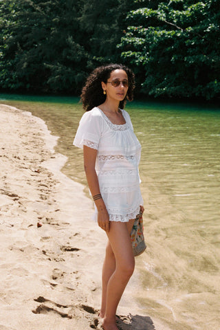 Woman in a white outfit standing on a sandy beach with greenery in the background