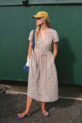 Woman in a floral dress and yellow cap standing against a green wall.
