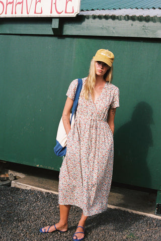Woman in a floral dress and yellow cap standing against a green wall.