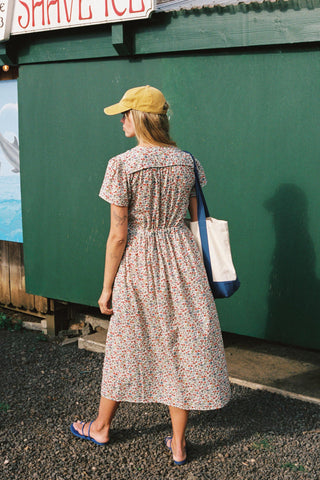 Woman in a floral dress and yellow cap walking away from a green wall.