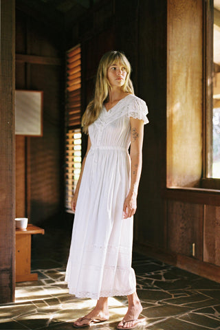 Woman in a white dress standing in a sunlit room with wooden walls.