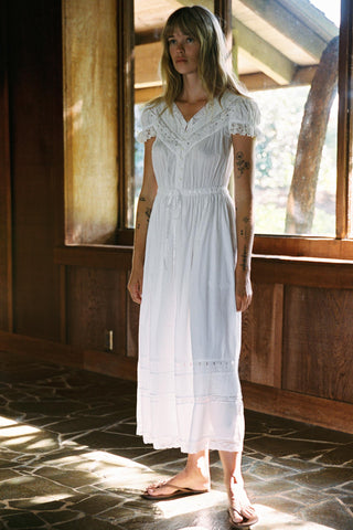 Woman in a white dress standing in a sunlit room with wooden walls.