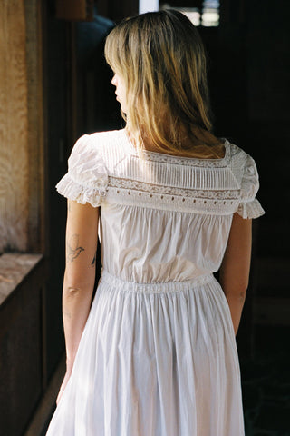 Woman wearing a white dress with lace details in a dimly lit room.