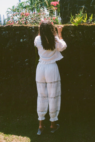 Person in a white outfit standing in front of a garden wall with plants and flowers.