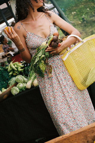 Woman in a floral dress holding vegetables and a yellow bag at an outdoor market.