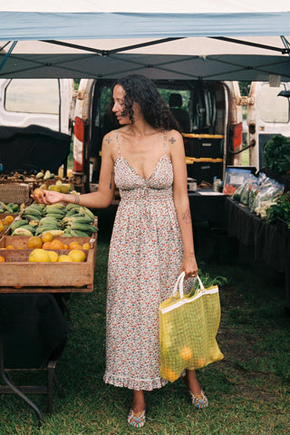 Woman in a floral dress holding a reusable bag at an outdoor farmers market.