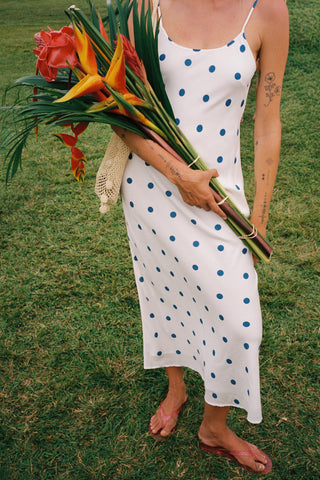 Person in a white dress with blue polka dots holding a bouquet of tropical flowers on grass.