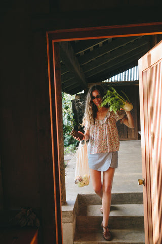 Woman holding a bunch of greens and a bottle, standing in a doorway with a garden view.
