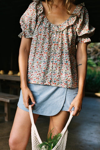 Woman wearing a floral blouse and blue skirt holding a string bag with groceries.