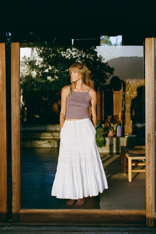 Woman standing in a doorway wearing a purple top and white skirt, with a scenic background.