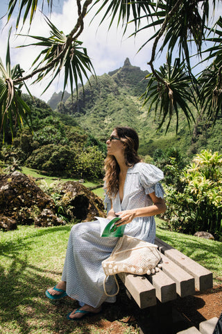 Woman in a dress sitting on a bench in a lush green garden with mountains in the background, with a woven bag on the bench