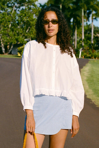 Woman wearing a white blouse and blue checkered skirt standing outdoors with greenery in the background