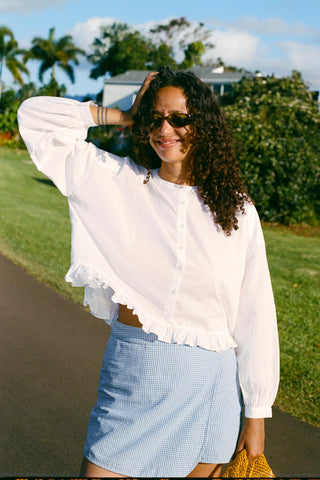 Woman in a white blouse and blue skirt standing outdoors with greenery in the background