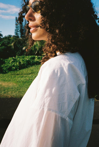 Woman wearing sunglasses and a white shirt outdoors with greenery in the background