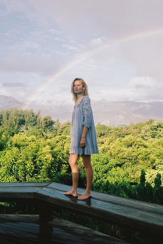 Woman wearing a light blue floral patterned dress standing on a wooden platform with a rainbow in the background