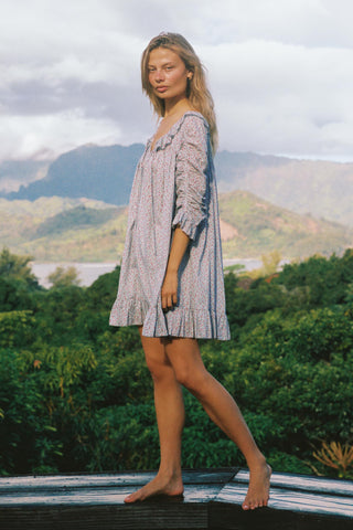 Woman wearing a light blue floral patterned dress standing outdoors with mountains and trees in the background