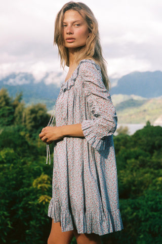 Woman wearing a light blue floral patterned dress standing in a scenic outdoor setting with mountains and trees.