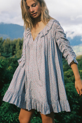 Woman wearing a light blue floral patterned dress standing in a natural setting with mountains and greenery.