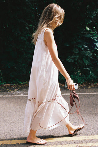 Woman in a white and embroidered dress walking outdoors with greenery in the background