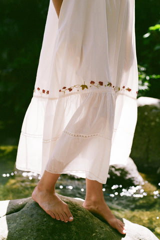 Person wearing a white dress with floral embroidery standing on a rock by water.