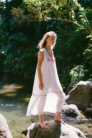 Woman in a white and embroidered dress standing by a stream with greenery in the background