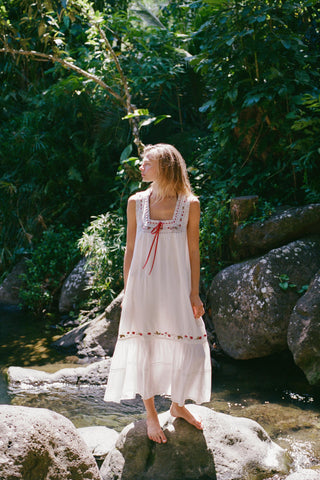 Woman in a white and embroidered dress  standing on rocks by a stream with greenery in the background