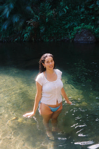 Woman in a floral top and blue bikini standing in clear water with greenery in the background