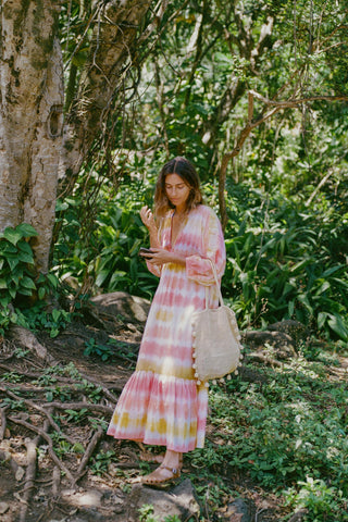 Woman in a pink and yellow tie-dyed dress standing in a forest with a woven bag
