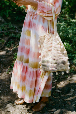 Person wearing a tie-dye dress holding a woven bag with pom-poms outdoors.