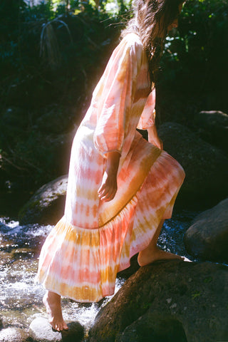 Person in a tie-dye dress standing by a stream with rocks and greenery.