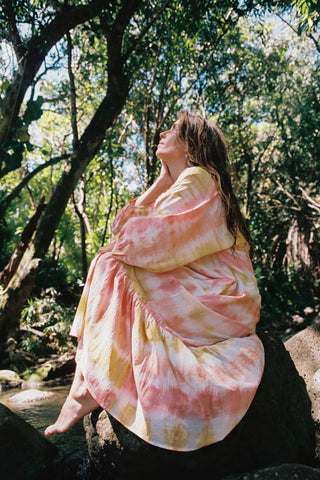 Person wearing a tie-dye dress sitting in a natural setting with trees and water.